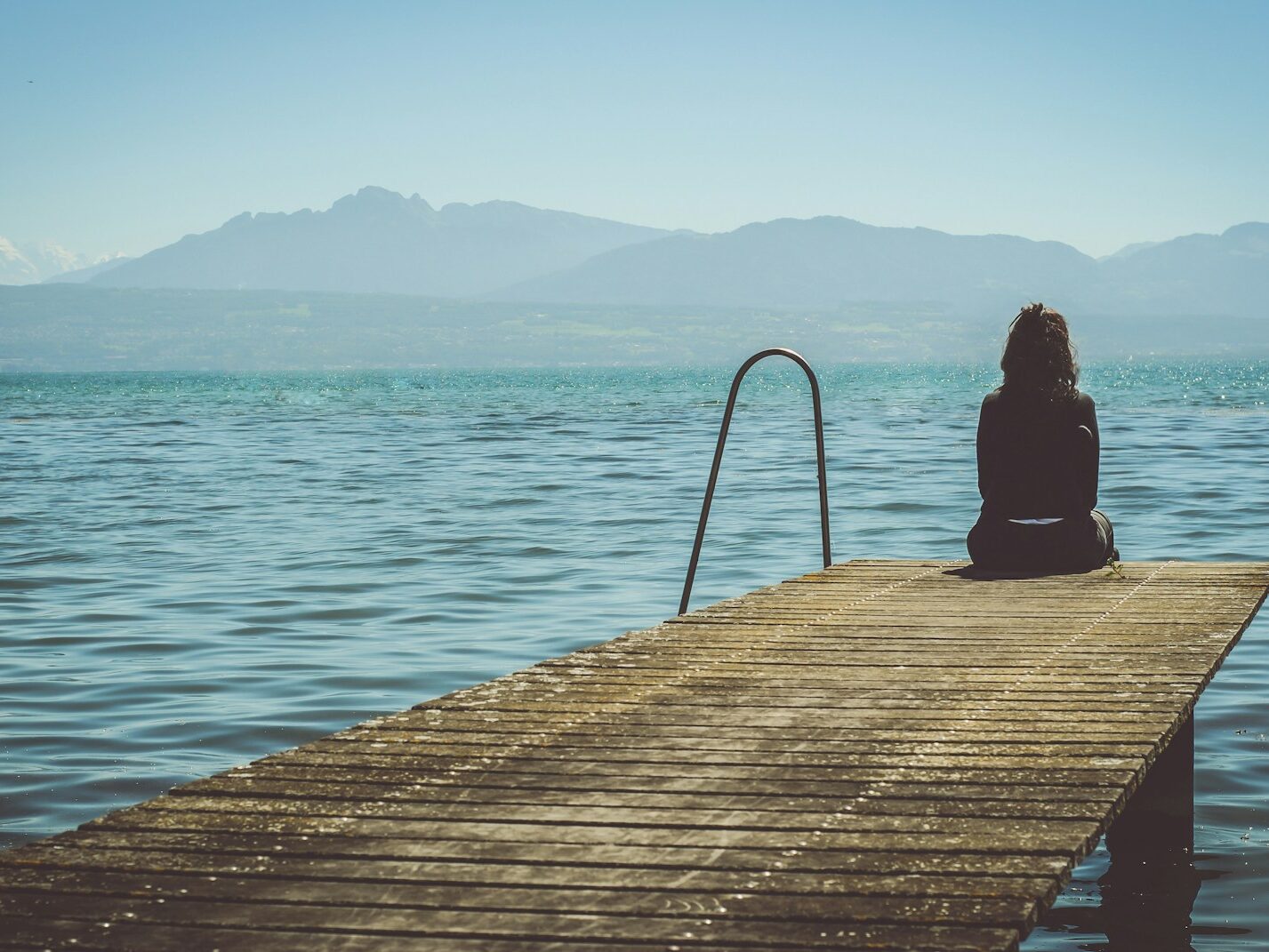 a woman sits on the end of a dock during daytime staring across a lake