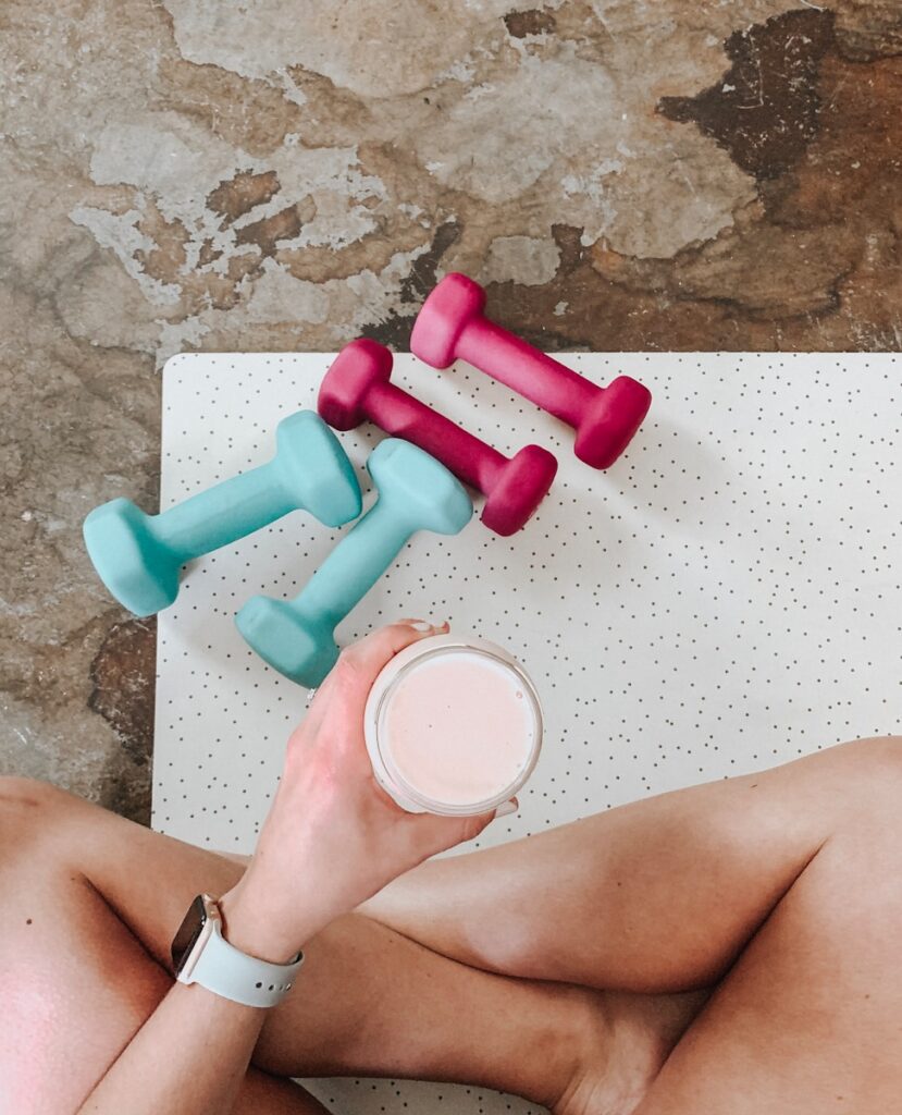 Woman holding protein shake on yoga mat beside small dumbbells—showing real-life fitness routines.