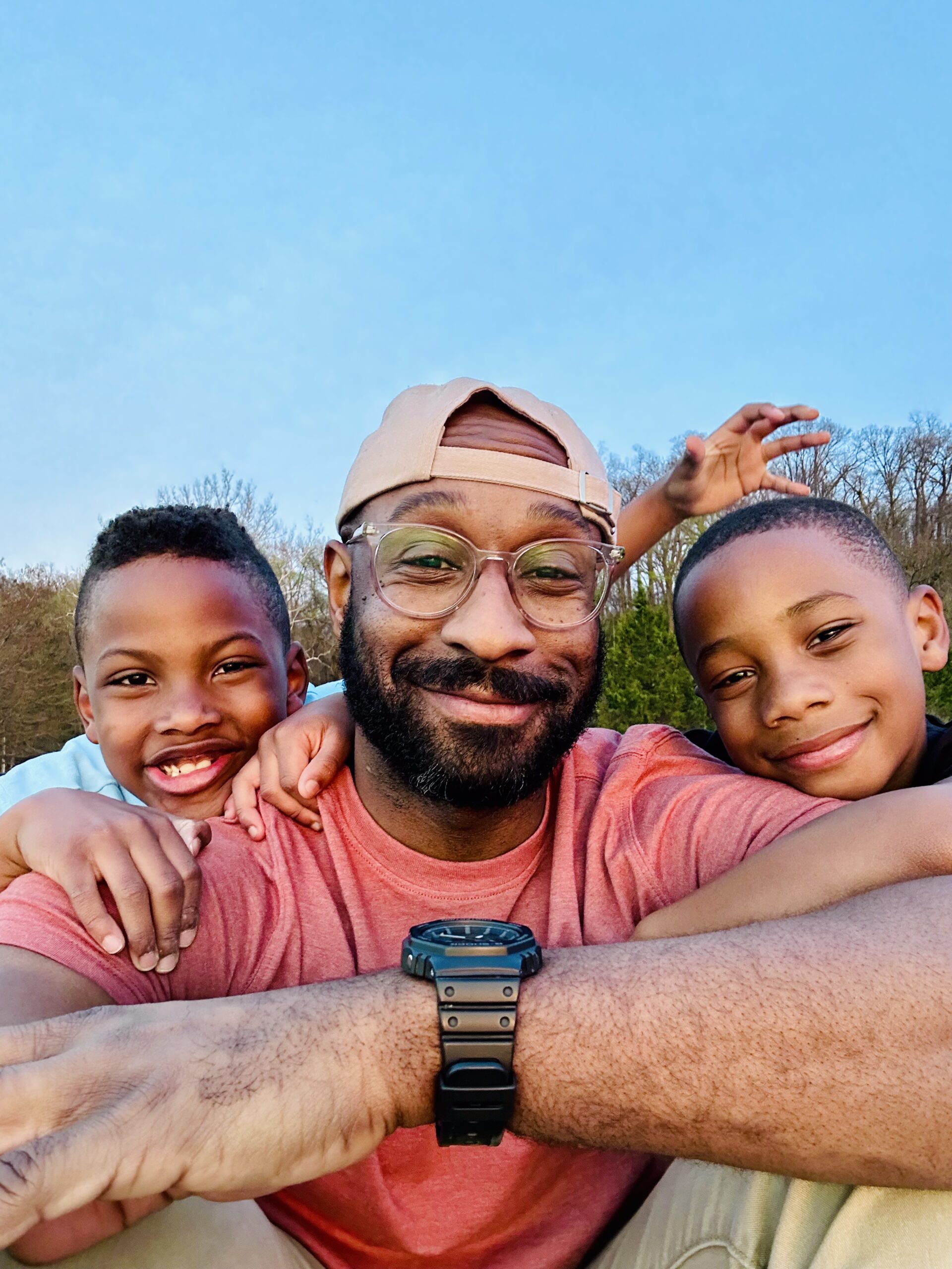 Justin smiling outdoors with his two young sons, all three with arms around each other and enjoying a sunny day.