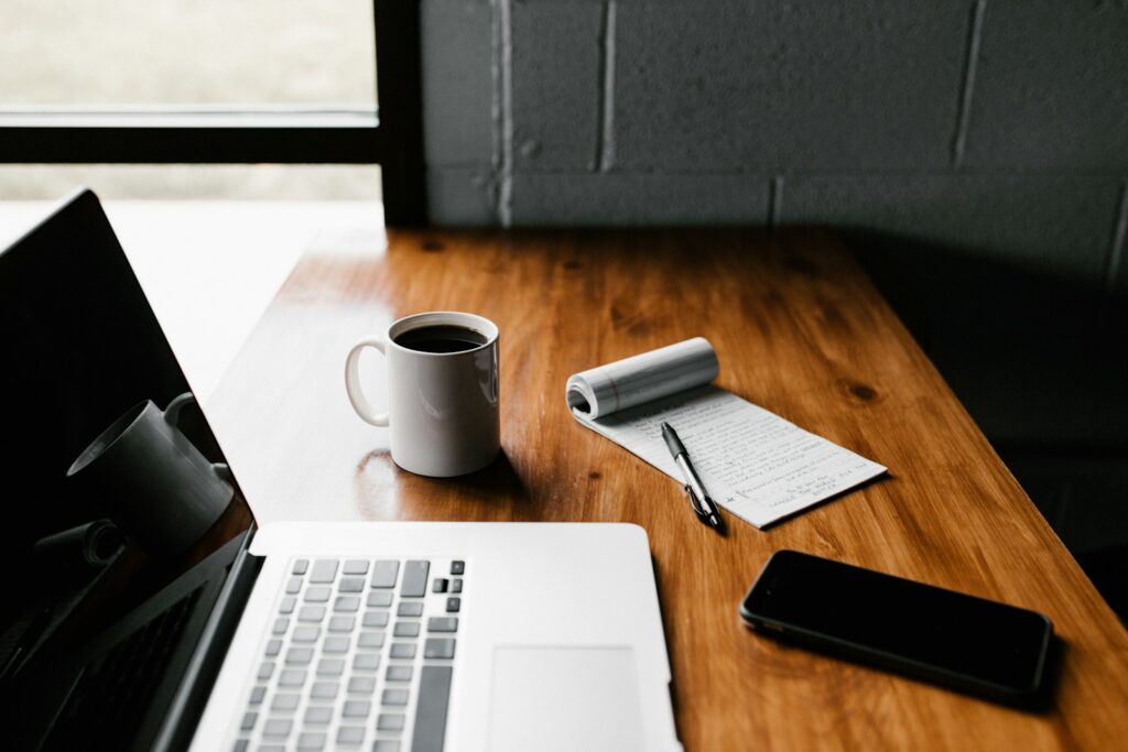 A clean, organized wooden desk featuring a white coffee mug, a laptop, a smartphone, and a notepad with a pen, conveying a sense of calm and intentionality.