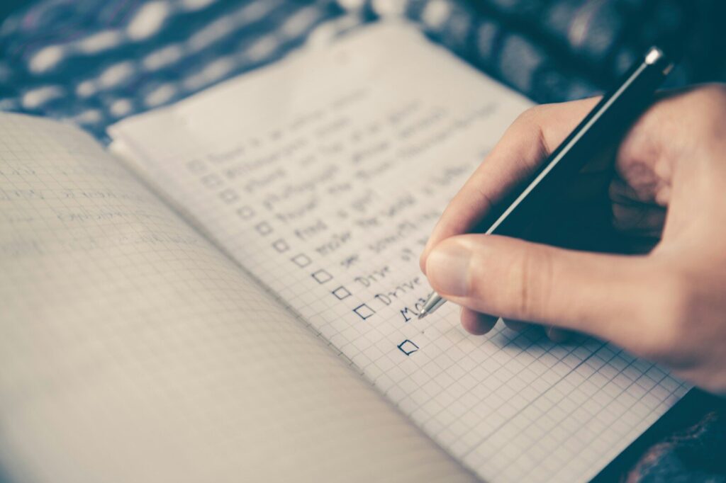 A close-up, slightly blurred photo of a person’s hand using a pen to check off items on a grid-lined notebook page, representing a focus on constant improvement and checklists.