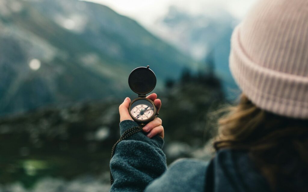 A person wearing a knit beanie holds an open brass compass in their hand, pointing toward a blurred mountain landscape in the distance.