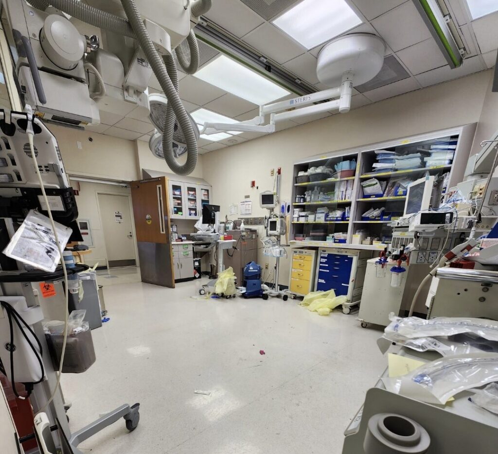 An empty trauma bay after a case, showing medical equipment, supply shelves, and scattered protective gear on the floor.