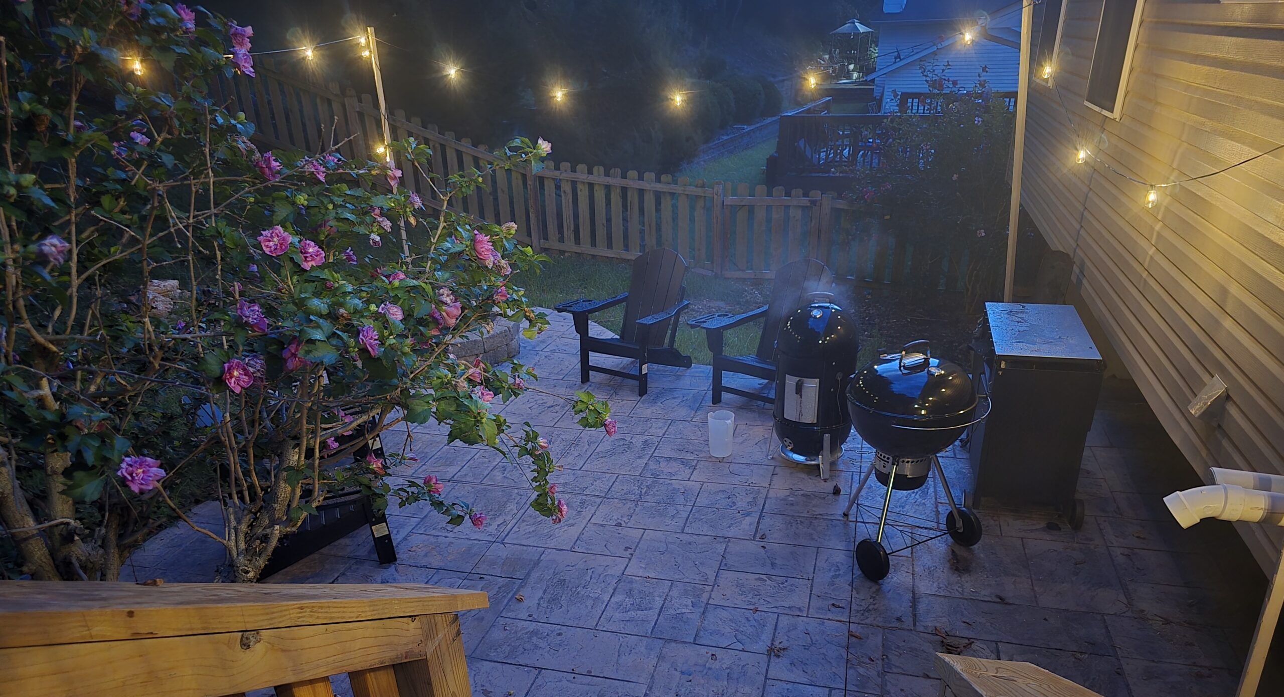 Evening view from a deck overlooking a backyard patio with string lights, flowers, and soft summer lighting.