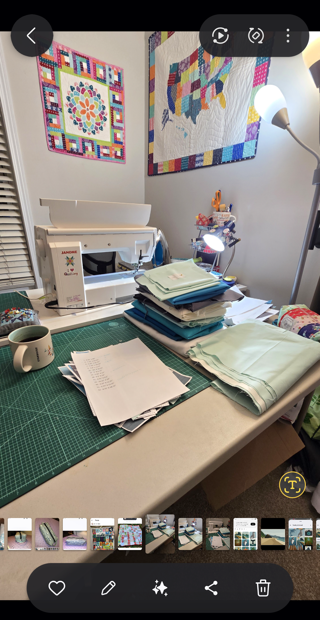 Sewing machine on a table with stacks of teal and mint fabric, printed pattern pages, a cutting mat and a mug of coffee, with two colorful quilts hanging on the wall above.